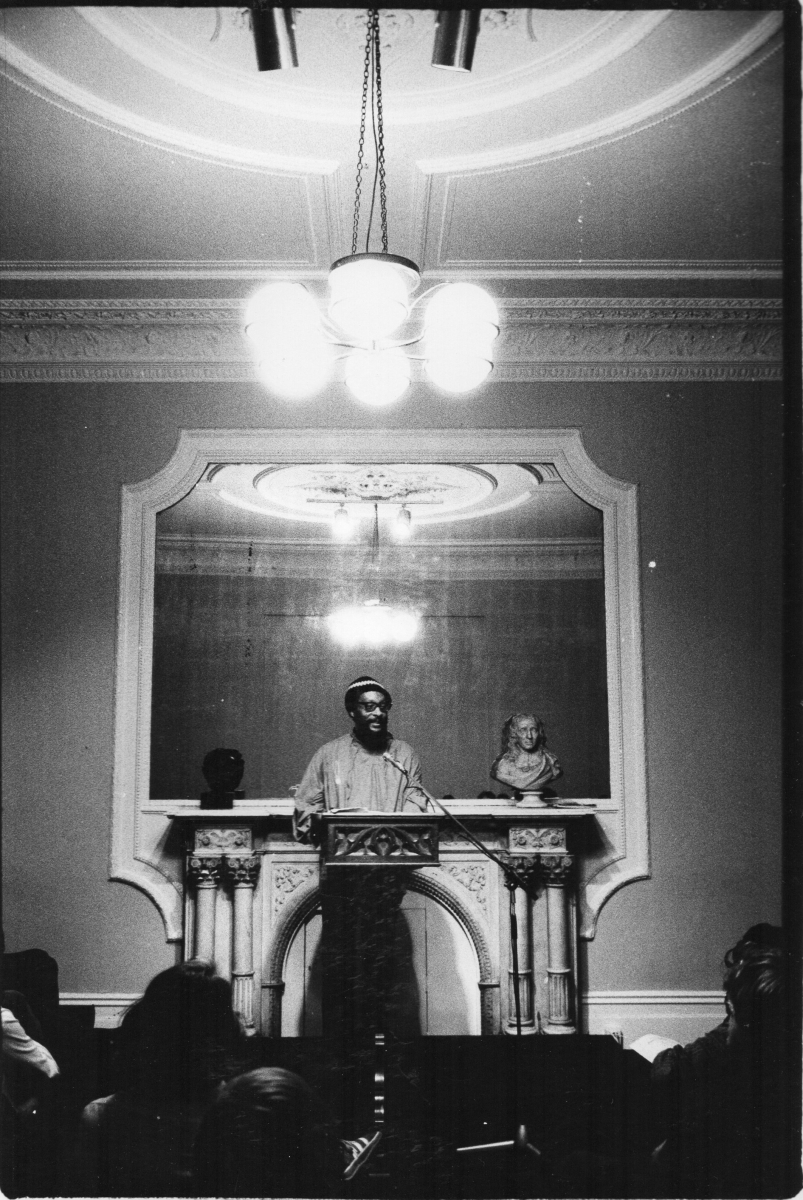 This photo is credited to the photographer George Hallett, based in France.  It shows Kamau reading at the Poetry Society in London, then at its home in Earl’s Court Square.  Again, Kamau has no memory of ever reading there, but Sarah White of NBB and I remember the event.  Poetry Society unable to help.  My 1989 diary records: “Wednesday 13 September, 7.30, EKB and Malik, Poetry Society.”  So suggest date is given as 1989.  (Kamau helpfully states that he did not begin to wear a tam until after 1986.)